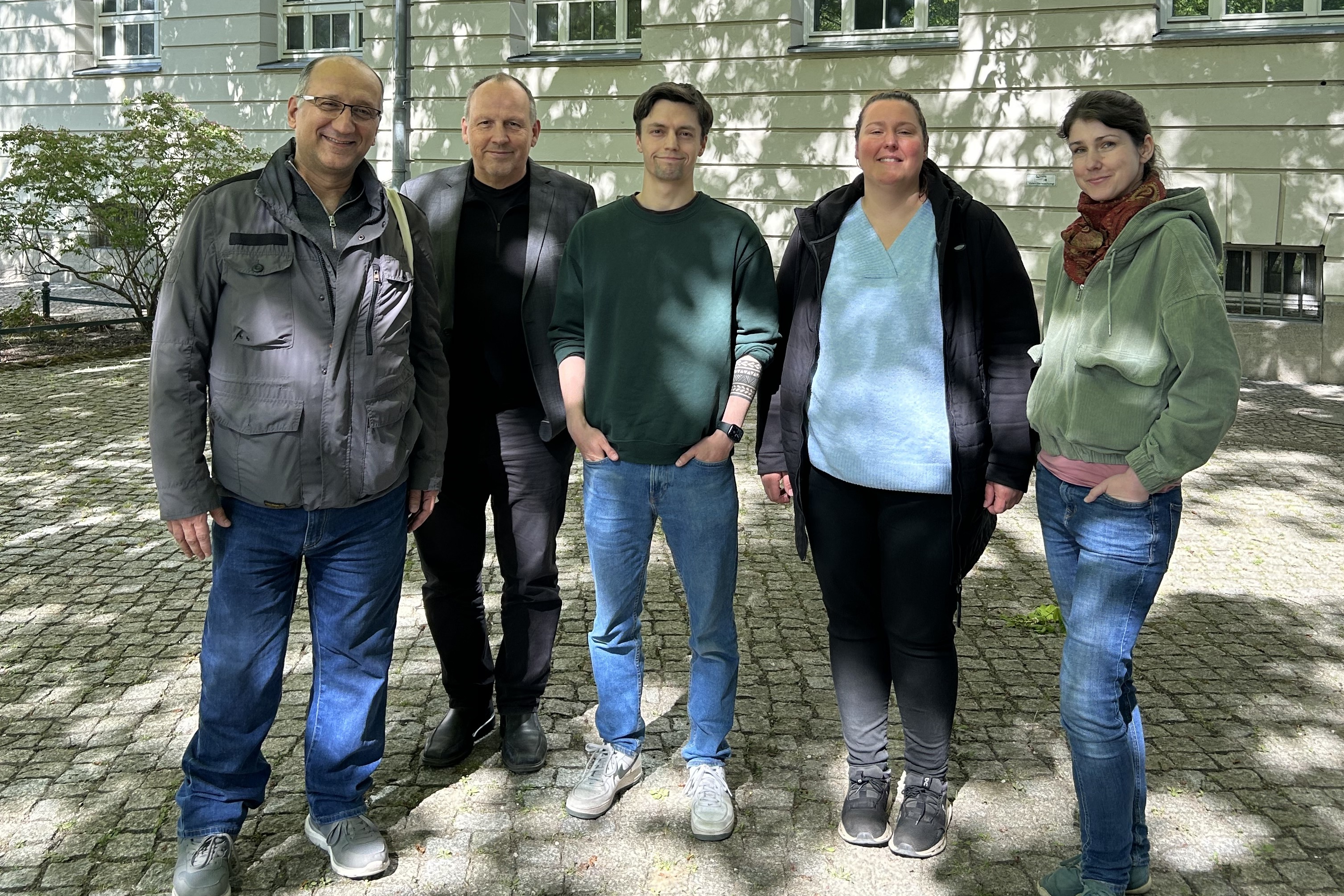Fünf Personen posieren vor einem Gebäude unter dem lichten Schatten eines Baumes