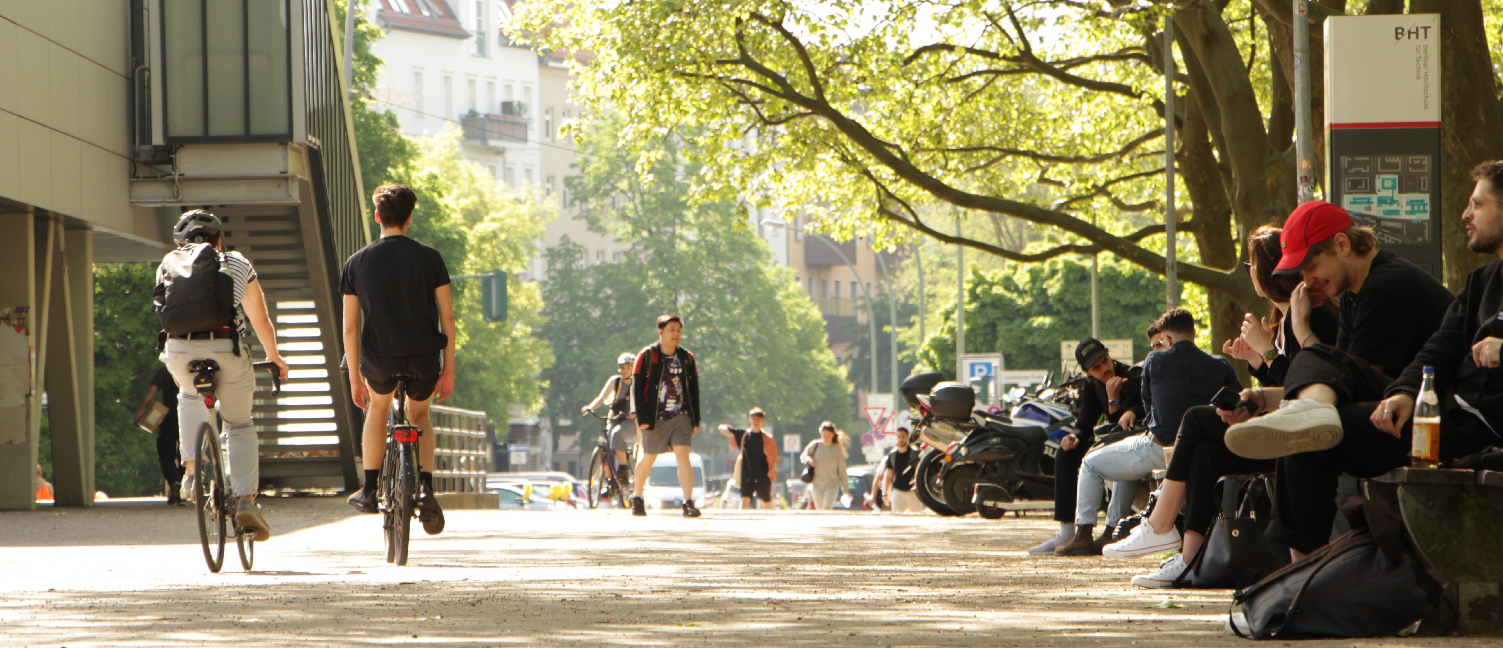 Menschen auf dem Campus der BHT, unter anderem zu Fuß und mit dem Rad unterwegs, im Hintergrund Autos und Motorräder 