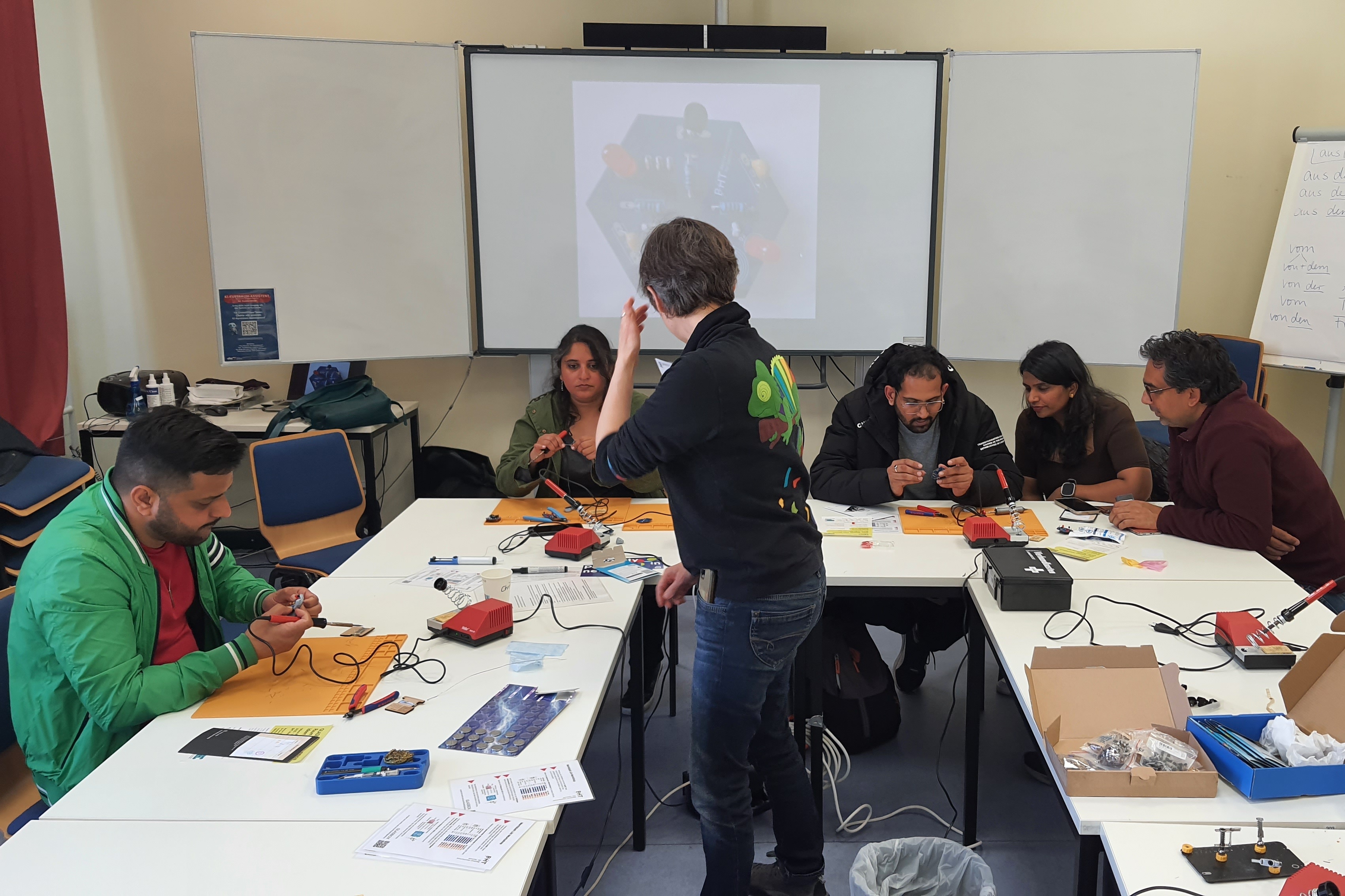 Mehrere Personen sitzen an Tischen eines Seminarraums mit Werkzeug und Materialen zum Löten