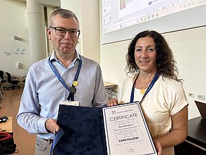 Ein Mann und eine Frau blicken in die Kamera, die Frau hält eine Urkunde