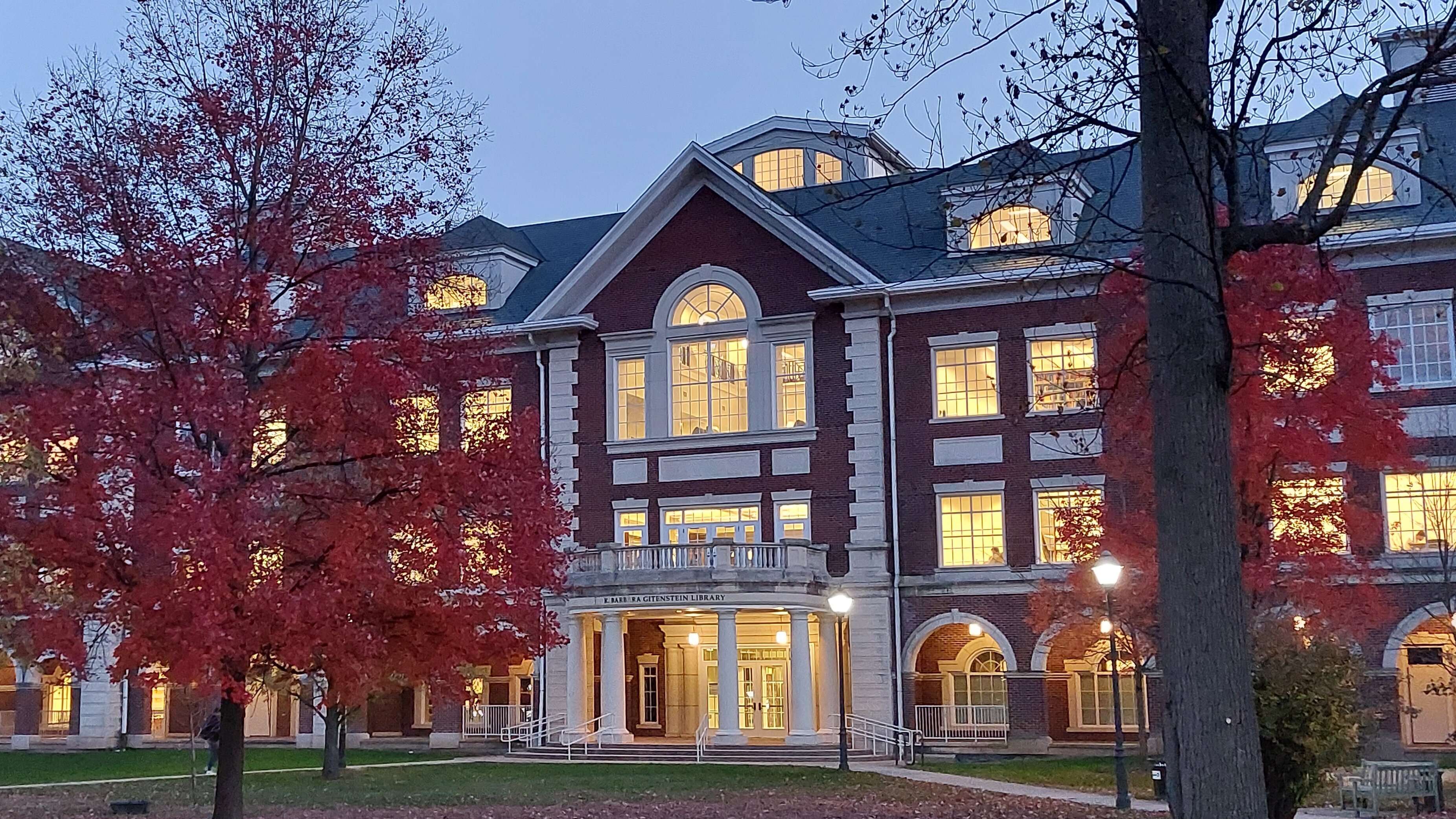Säuleneingang eines klassischen Uni-Gebäudes in der Dämmerung, Licht strahlt durch Fenster, davon Rot-belaubte Bäume