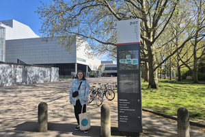 Eine junge Frau mit hellblauer Jacke, dunkler Hose und Jutebeutel der Berliner Hochschule für Technik (BHT) steht neben einem BHT Wegweiser mit BHT Lageplan und verschiedenen Richtungsangaben. Im Hintergrund sieht man das BHT Gebäude Haus Bauwesen.