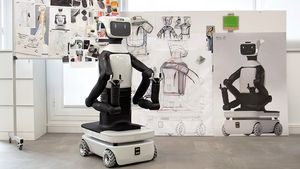 Foto eines schwarz-weißen humanoiden Roboters: Sein Unterteil ist wie ein kleines Auto, vom rechteckigen Korpus gehen zwei Arme mit Klauen vorn ab. Oben sitzt ein kleiner Bildschirm mit zwei Augen und einem lächelnden Mund. Hinter ihm hängen an einem Flipchart Konzeptzeichnungen weiterer Roboter.