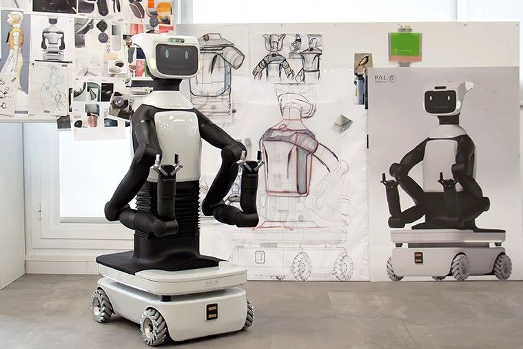 Foto eines schwarz-weißen humanoiden Roboters: Sein Unterteil ist wie ein kleines Auto, vom rechteckigen Korpus gehen zwei Arme mit Klauen vorn ab. Oben sitzt ein kleiner Bildschirm mit zwei Augen und einem lächelnden Mund. Hinter ihm hängen an einem Flipchart Konzeptzeichnungen weiterer Roboter.