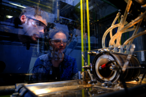 Ein Student und eine Studentin betrachten einen technischen Gegenstand durch eine Glasscheibe hindurch.