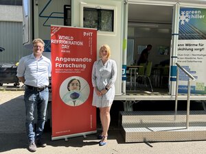 Ein Mann und eine Frau stehen vor einem Trailer, zwischen Ihnen ein Aufsteller mi der Aufschrift: Angewandte Forschung