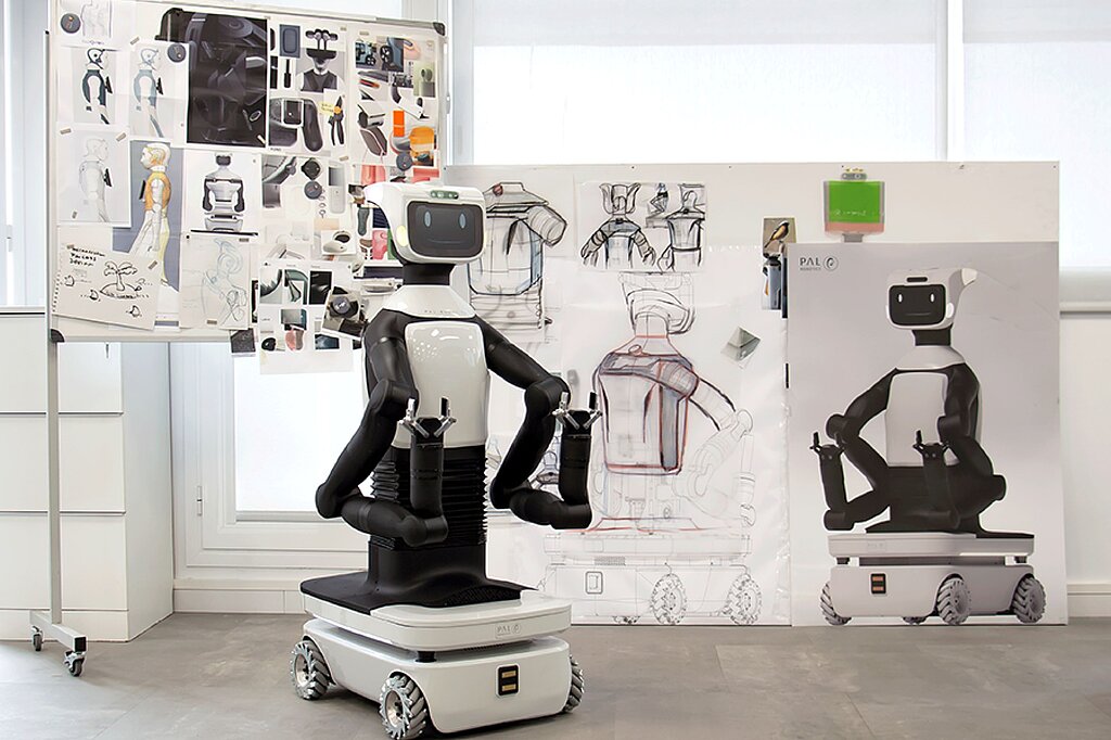 Foto eines schwarz-weißen humanoiden Roboters: Sein Unterteil ist wie ein kleines Auto, vom rechteckigen Korpus gehen zwei Arme mit Klauen vorn ab. Oben sitzt ein kleiner Bildschirm mit zwei Augen und einem lächelnden Mund. Hinter ihm hängen an einem Flipchart Konzeptzeichnungen weiterer Roboter.
