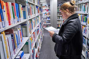 Eine Person steht zwischen Buchregalen in der Bibliothek und sieht sich ein Buch an.