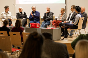 Acht Personen sitzen auf einem Podium vor gefüllten Stuhlreihen