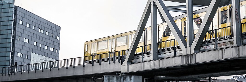 ReComTrans Brücke, auf der eine gelbe U-Bahn fährt. Dahinter ein modernes Hochhaus.