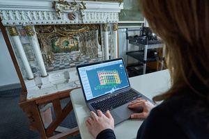 Eine Frau arbeitet am Laptop an einem digitalen Bühnenmodell, davor ist ein plastisches Modell einer Theaterbühne zu sehen