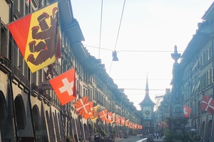 Blick in eine mit Schweizer und Berner Flaggen geschmückte Straße in Bern - rechts im Bild ein dekorativer Brunnen, im Hintergrund ein Turm mit großer Uhr.