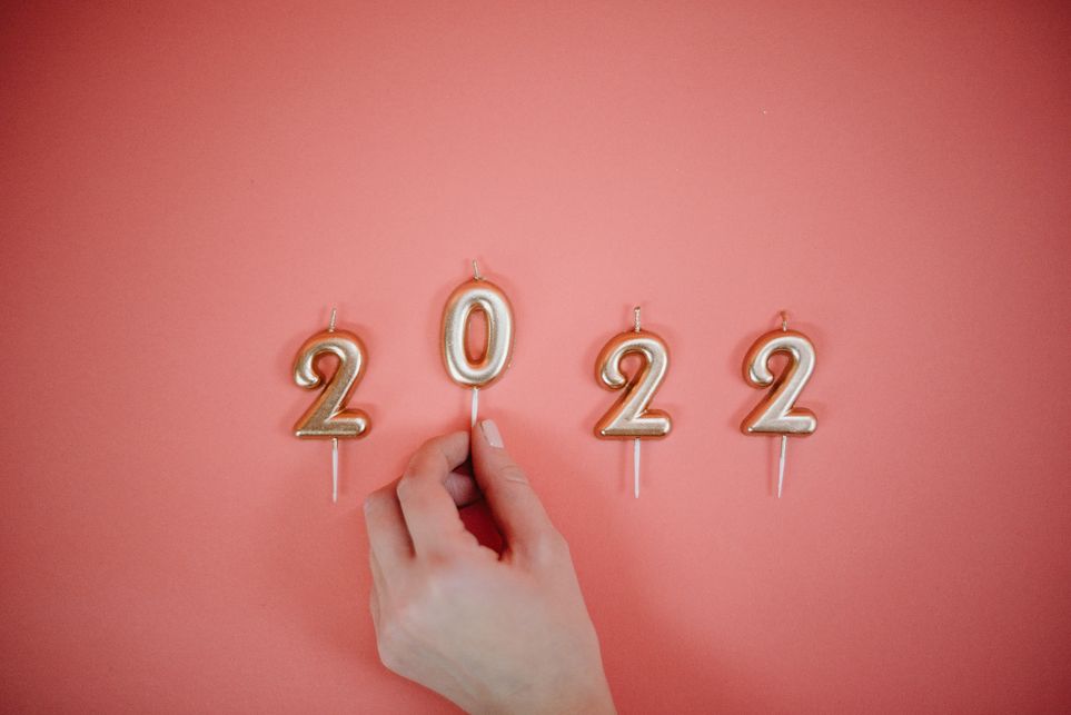 Das Bild zeigt eine Hand vor einem rosafarbenen Hintergrund. Die Hand legt gerade eine Kerze in Form einer goldenen Null hin, auf beiden Seiten befinden sich dementsprechend weitere Kerzen in Form von goldenen Zweien. Diese Kerzen bilden 2022 zum Auftakt des neuen Jahres.