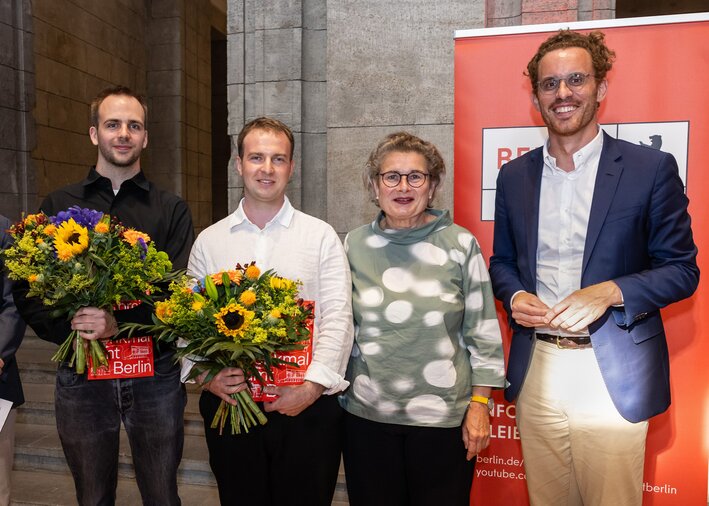 Vier Personen stehen nebeneinander und blicken in die Kamera, der erste und der zweite von Links halten Blumensträuße in den Händen