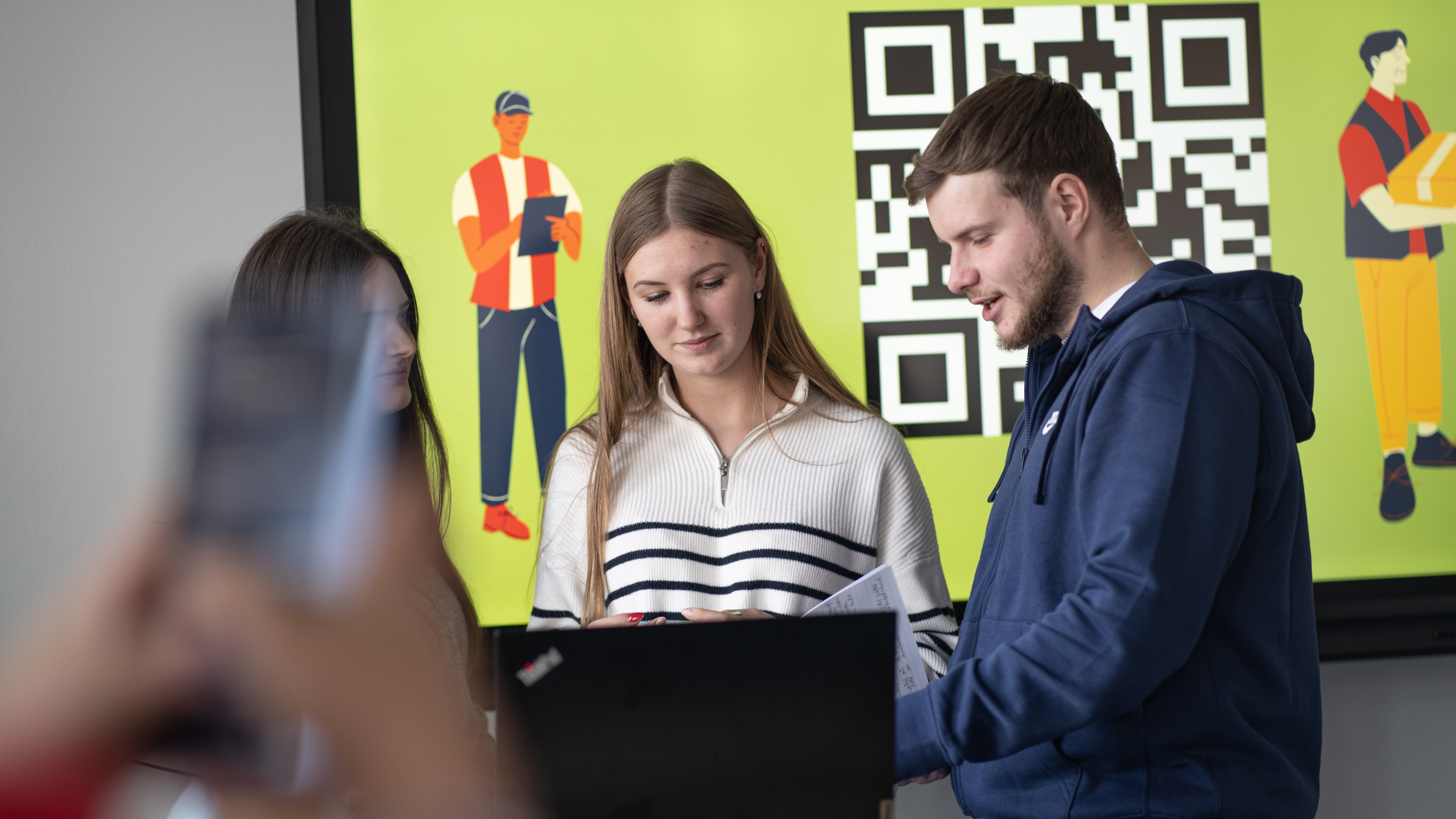 Drei junge Menschen stehen um einem Laptop, hinter Ihnen ist eine Präsentation mit großem QR-Code zu sehen