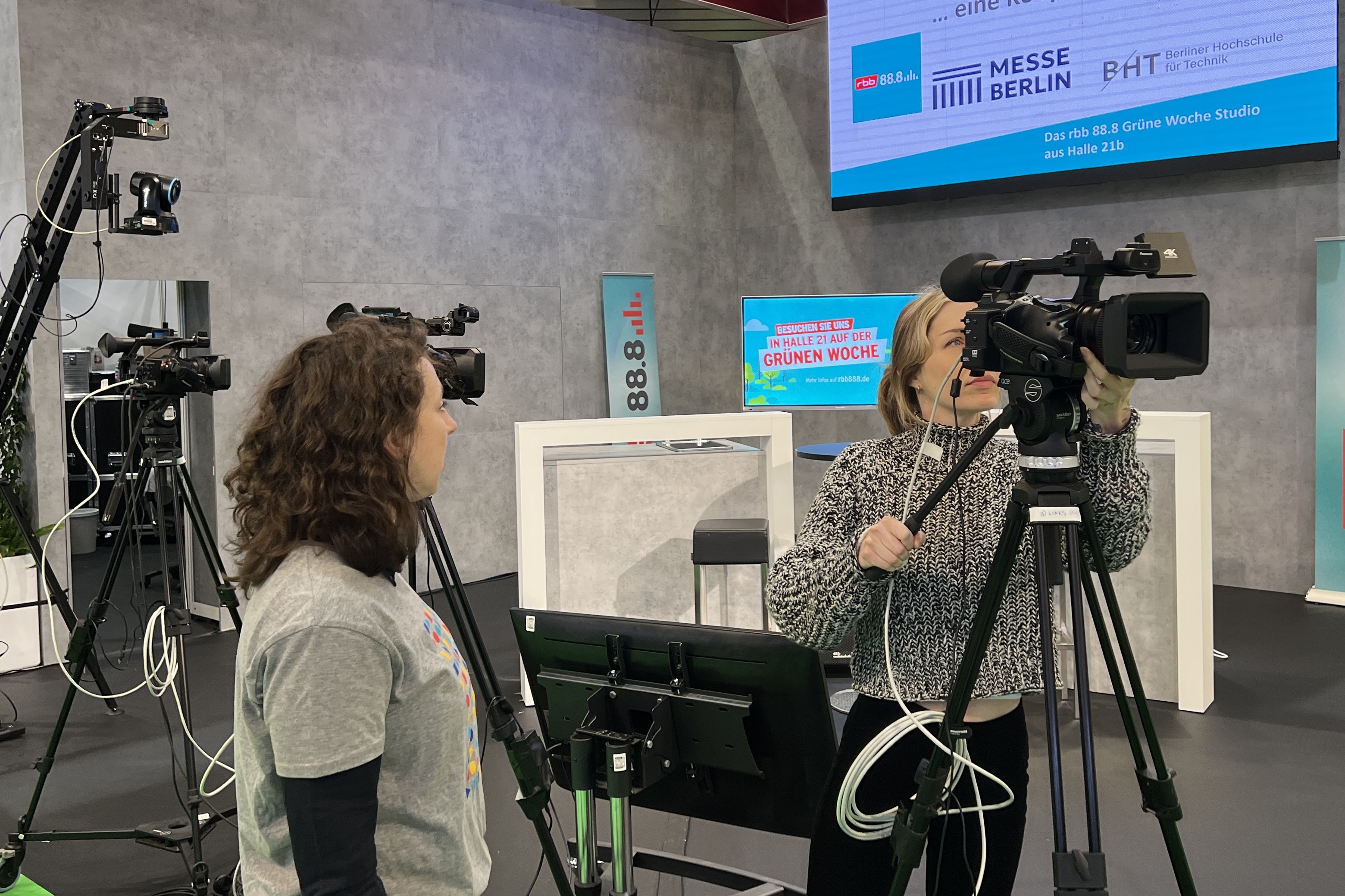 Vor einem Messerstand blickt eine Frau in eine Filmkamera, eine weitere Frau steht daneben und blickt zu ihr