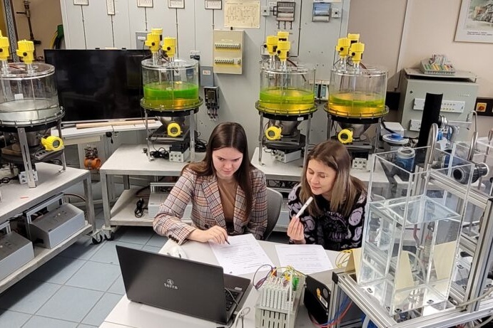 Zwei Personen sitzen in einem Labor vor einem Laptop. Zahlreiche Apparaturen und Gefäße mit Flüssigkeiten sind zu sehen.
