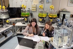 Zwei Personen sitzen in einem Labor vor einem Laptop. Zahlreiche Apparaturen und Gefäße mit Flüssigkeiten sind zu sehen.