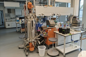 Blick auf den Bioreaktor Labor für Umwelt- und Bioverfahrenstechnik: Ein Edelstahlzylinder, aus dem verschiedene Schläuche hinaus- und wieder hineinführen, mit einem Auslassventil und einer Bedienstation. Daneben ein Labortisch mit drei großen Flaschen voll dunkler Flüssigkeit, die in den Reaktor hineinläuft.