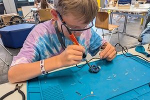 Junge mit Schutzbrille in Batik-Shirt lötet etwas auf einen Platine in einem Lehr-Labor