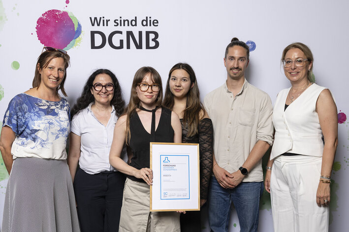 Sechs Personen posieren für ein Foto, die Zweite von Links hält eine gerahmte Urkunde in der Hand