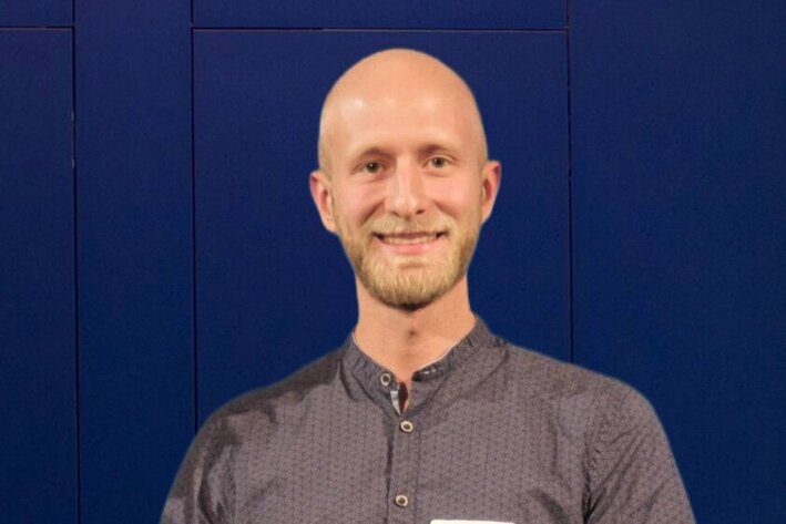 Mann hält eine aufgeklappte Urkundenmappe vor sich und steht vor einer großen blauen Tür