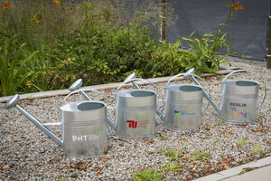 Vier Metallgießkannen vor einem Beet mit Logos von BHT, TU Berlin, Tegel Projekt und Berliner Wasserbetrieben