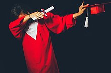 Promotion Eine Person mit rotem Abschlusskleid hält einen roten Promotionshut und eine Schriftrolle in der Hand