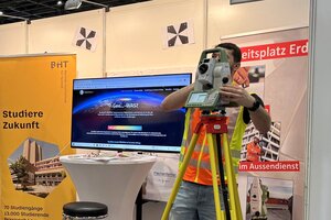 Vor einem Messestand blickt ein Student durch ein Vermessungsgerät, ein anderer trägt eine VR-Brille