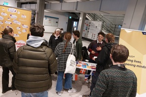 Drei Personen befinden sich hinter einem Tisch mit Flyern und Länderfahnen und scheinen junge Menschen zu beraten, die um den Tisch herumstehen. Neben dem Tisch sind gelbe Roll-Ups von Erasmus+ und Studieren Weltweit.