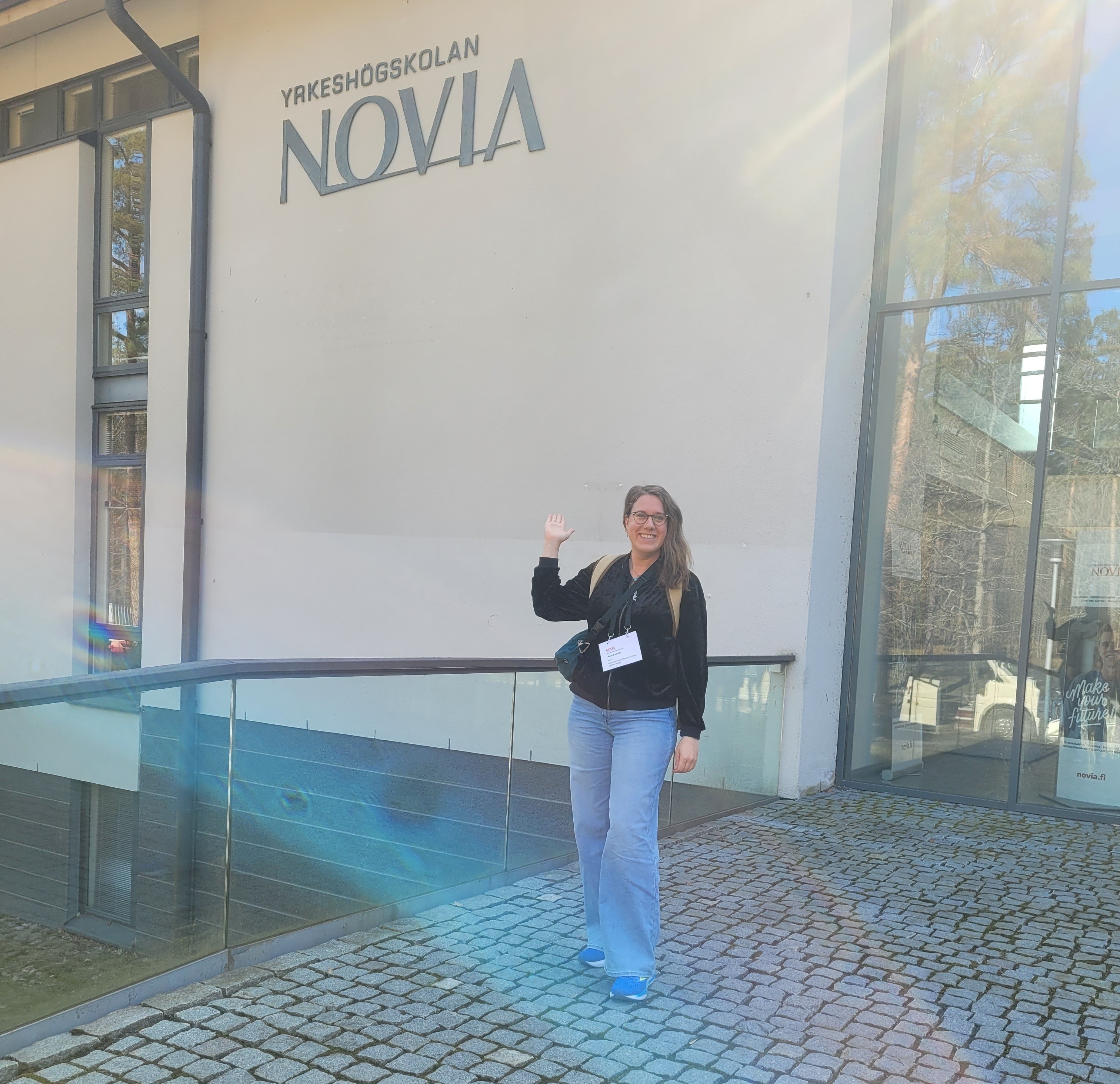 Eine junge Frau mit hellblauer Jeans, schwarzem Oberteil, Rucksack und Umhängetasche steht lächelnd auf Kopfsteinpflaster vor einem Gebäude und zeigt auf den Schriftzug "Yrkeshögskolan NOVIA".