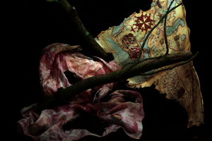 Teile einer historischen Weltkarte und rosafarbener Seidenstoff sind an Ästen mit Seidenfäden versponnen. Der Hintergrund ist ein erdiges Schwarz.