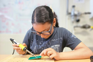 Junge Frau sitzt an einem Tisch und lötet etwas auf eine Platine