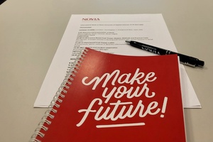 Ein roter Notizblock mit der Aufschrift "Make your Future" liegt mit einem Kugelschreiber und Papier auf einem Schreibtisch. Im Hintergrund ist ein Bildschirm mit verschwommener Schrift zu sehen.
