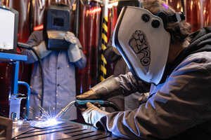 Eine Person mit Sicherheitskleidung und Maske schweißt, einen weitere Person mit Sicherheitsausrüstung im Hintergrund