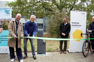 Mehrere Personen stehen vor einem Baum, dazwischen Roll ups und ein grauer Kasten. Zwei Menschen halten und schneiden ein grünes Band durch.