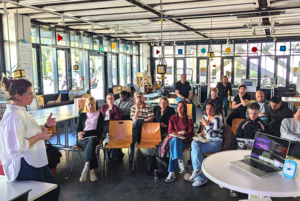 In einem hellen Raum steht eine Person vor einer Gruppe Menschen, die auf Stühlen sitzen.