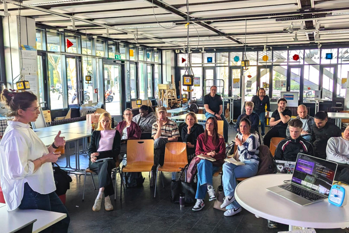 In einem hellen Raum steht eine Person vor einer Gruppe Menschen, die auf Stühlen sitzen.