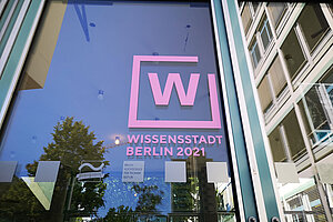 Eingang des Hauses Grashof mit Aufkleber "Wissensstadt", in der Glastür spiegelt sich der Campus