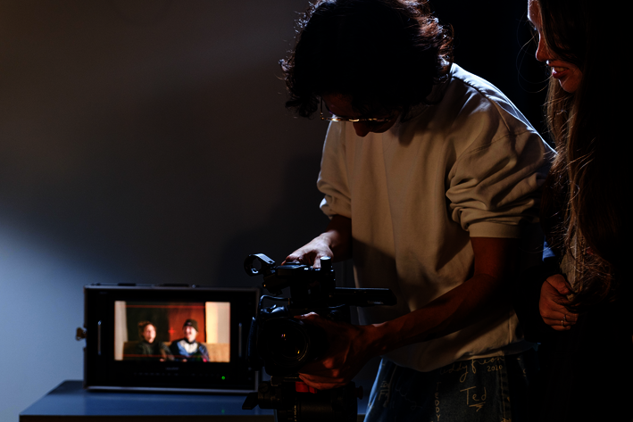 Zwei Personen halten eine Kamera und schauen auf diese. Im Hintergrund ist ein Monitor zu sehen.