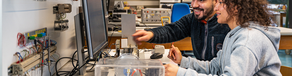 Ein junger Mann und eine junge Frau schauen auf einen Bildschirm. Auf dem Tisch vor dem Bildschirm sowie an den Wänden und auf einem weieren Tisch im Raum sind Kabel, Prüfgeräte und weitere Ausstattungsgegenstände des elektrotechnischen Labors zu sehen.