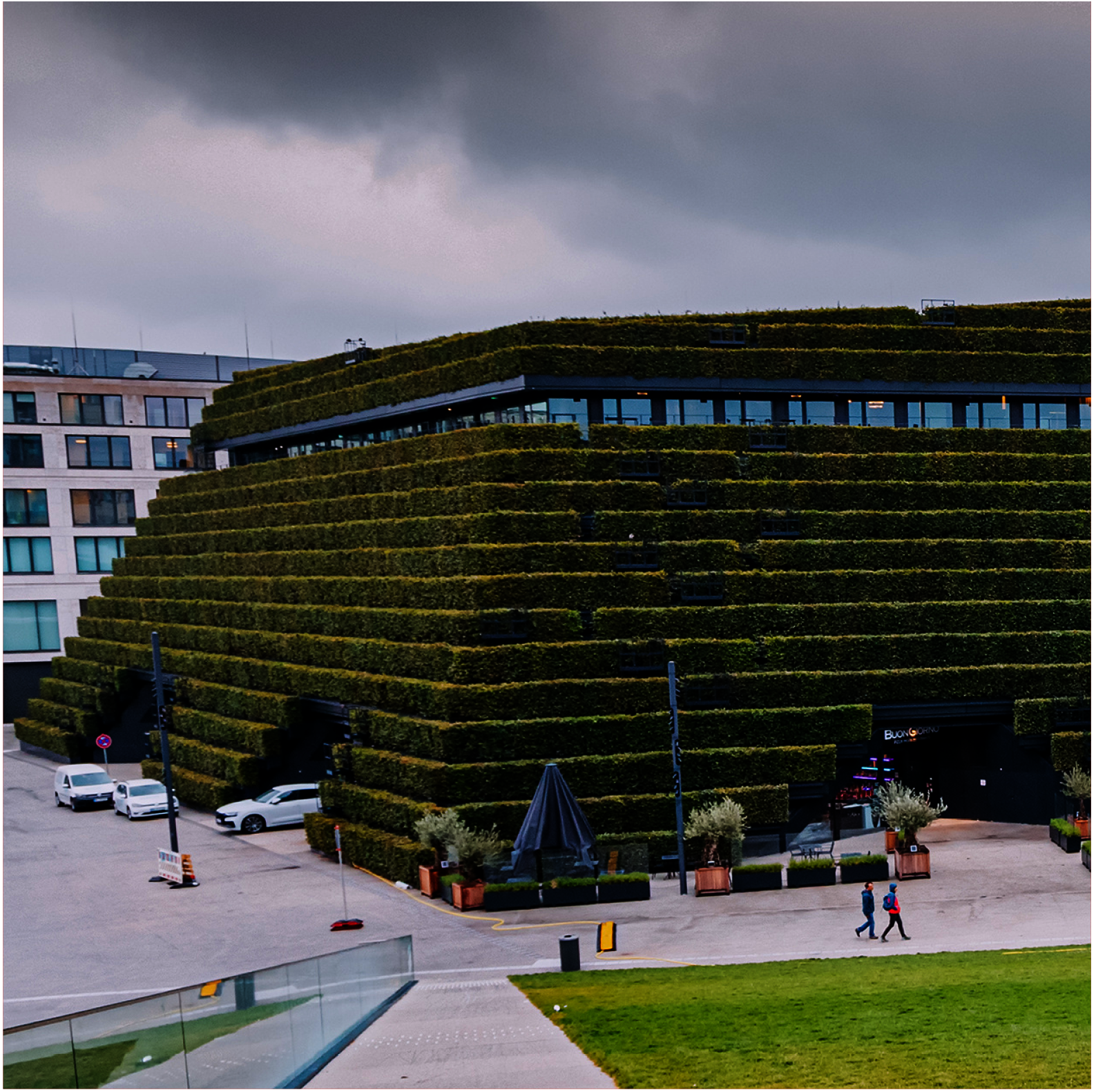 Blick aus der Vogelperspektive auf ein Gebäude mit schräger und in Stufen umlaufender Fassade, die komplett begrünt ist. Davor eine Grünfläche und Menschen, rechts reicht der Blick auf eine rechst um die Kurve laufende Straße