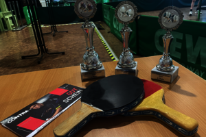 Auf einem Tisch befinden sich zwei Tischtennisschläger und drei Pokale.