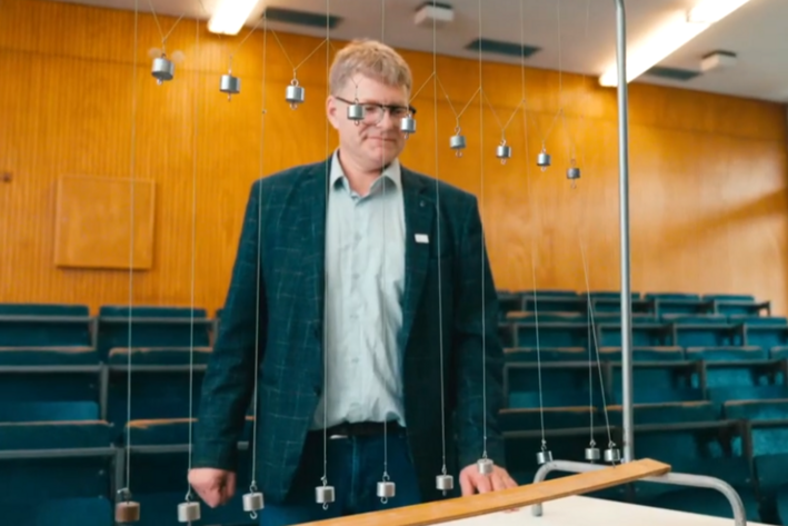 Andreas Modler steht in einem Hörsaal an einer Apparatur mit mehreren aufgereihten Pendeln