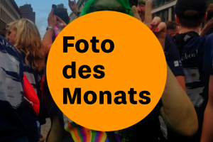 Bildaufschrift: „Foto des Monats“. Im Hintergrund ist eine Menschenmenge zu sehen.