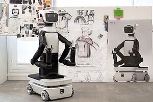 Foto eines schwarz-weißen humanoiden Roboters: Sein Unterteil ist wie ein kleines Auto, vom rechteckigen Korpus gehen zwei Arme mit Klauen vorn ab. Oben sitzt ein kleiner Bildschirm mit zwei Augen und einem lächelnden Mund. Hinter ihm hängen an einem Flipchart Konzeptzeichnungen weiterer Roboter.