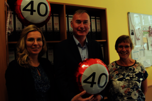 Drei Personen in einem Büro, der Mann in der Mitte hält einen Luftballon mit der Aufschrift „40“.