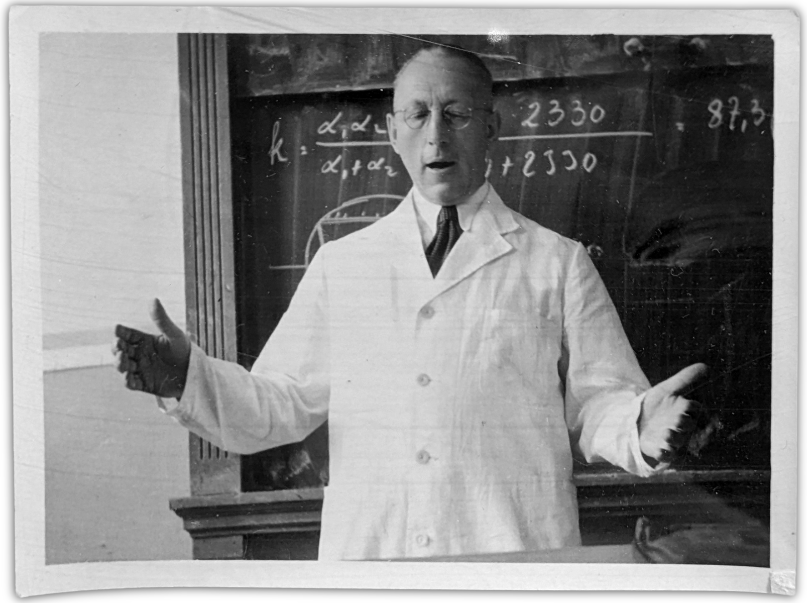 Schwarz-Weiß-Fotografie: Mann in weißem Kittel, darunter Hemd und Krawatte vor einer Tafel mit mathematischen Formeln, der den Zuhörenden etwas erklärt