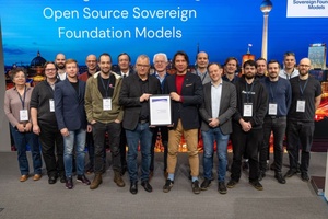 Eine große Personengruppe steht vor einer Wand, auf der Open Source Sovereign Foundation Models geschrieben steht. Zwei Personen stehen in der vordersten Reihe und halten gemeinsam ein gerahmtes Dokument in die Kamera.