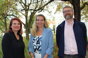 Drei Personen stehen vor grünen Bäumen und blicken freudig in die Kamera